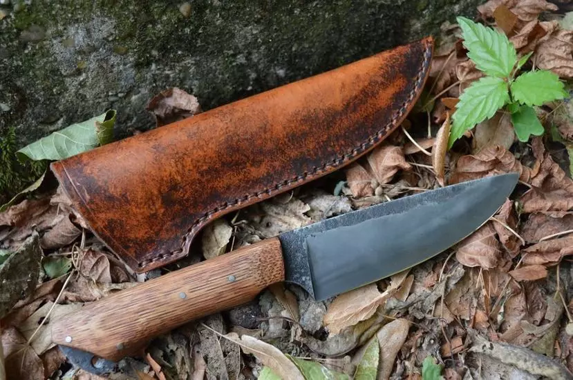 un couteau avec manche en bois et lame d'acier et à côté un fourreau en cuir ; le tout posé sur des feuilles