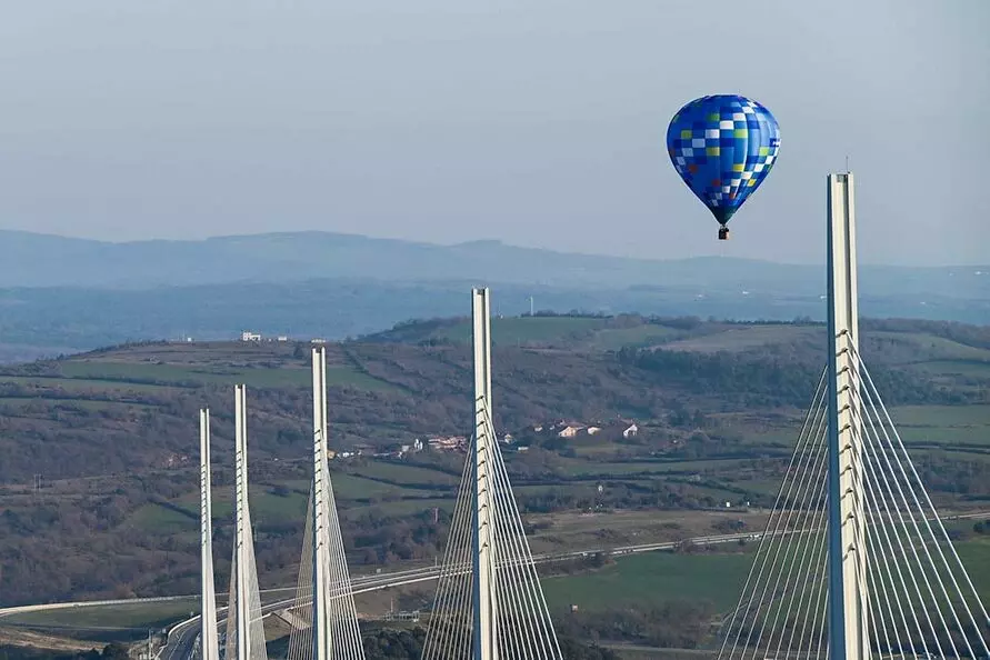 une montgolfière bleue survole le viaduc de millau ; au fond de la vallée, routes et autoroute ; en arrière plan, les monts de l'aveyron
