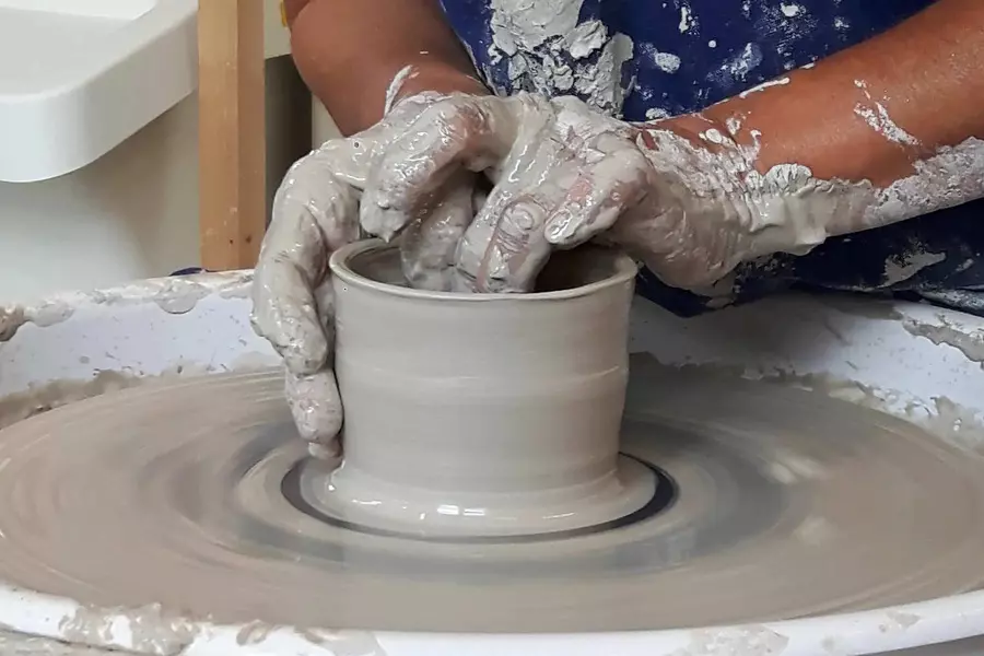 les mains d'une femme font lentement s'élever une poterie faite au tour tandis que celui-ci tourne ; la potière a les mains couvertes de glaise.