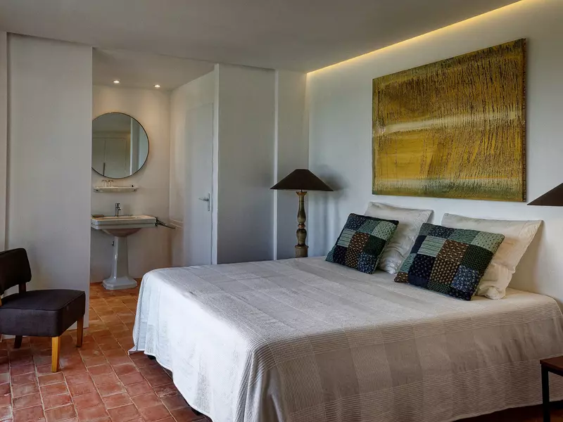 chambre du rez-de chaussée du gite la cubane près des gorges de l'aveyron en occitanie ; un lit recouvert de son plaid gris, des oreillers en mosaïque de bleu, gris et marron, au fond le lavabo ; au mur un tableau contemporain