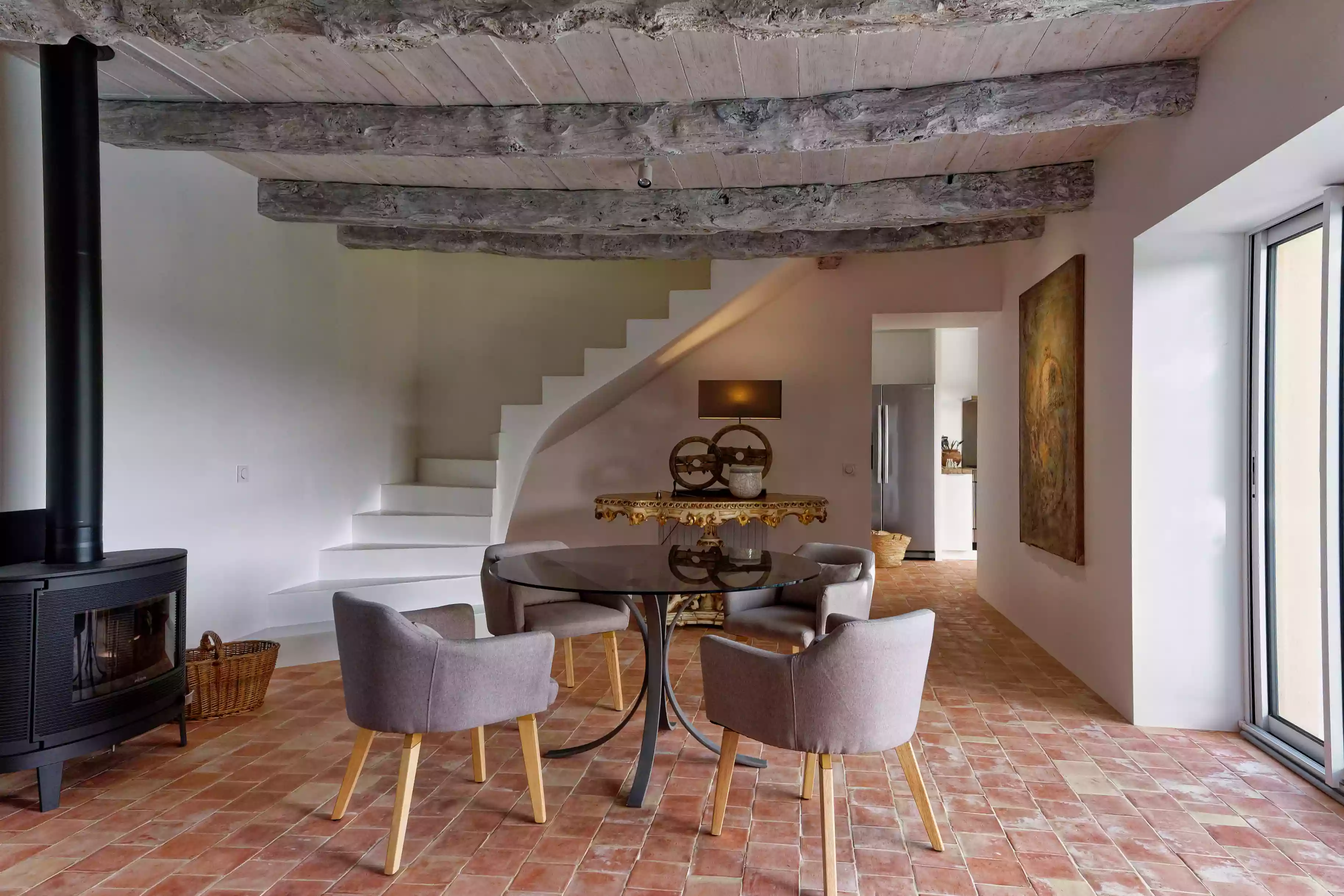 le salon-salle à manger du gîte de la cubane, avec le contraste entre un sol en tommettes et un plafond à poutre d'aspect traditionnel et une décoration résolument contemporaine ; au fond, la cuisine ; sur le côté, le poêle à bois