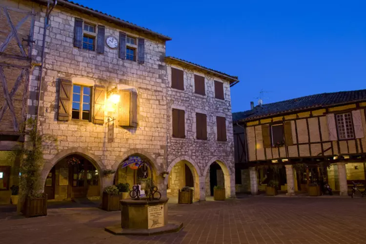 vue de la grand place de Castelnau Montmirail (tarn, occitanie) ; photo de nuit avec éclairage, au centre la fontaine ; les maisons sont à arcades, en pierre du pays ou en brique et colombage