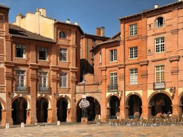 un angle de la place nationale à Montauban : les maisons sont en brique, avec des arcades en rez de chaussée ; il y a une terrasse de café sur la place