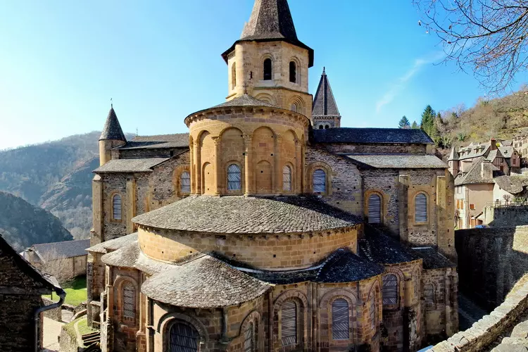 l'extérieur de l'église sainte foy de conques dans l'aveyron ; la photo montre l'abside avec ses toitures étagées en lauze ; le tout est coiffé par le clocher