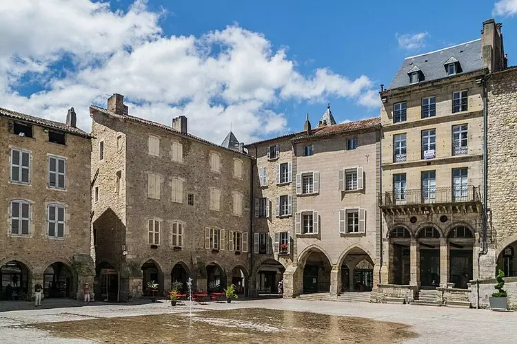 la place notre dame de villefranche de rouergue en aveyron, occitanie ; on voit les maisons en pierre avec les arcades en rez-de-chaussée ; la place est humide, la fontaine contemporaine venant juste de s'éteindre