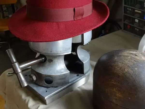 une atelier de chapellerie, avec un chapeau en feutre sur une presse pour lui donner la forme ; le chapeau est rouge, il est terminé et a été entouré d'un ruban, rouge également
